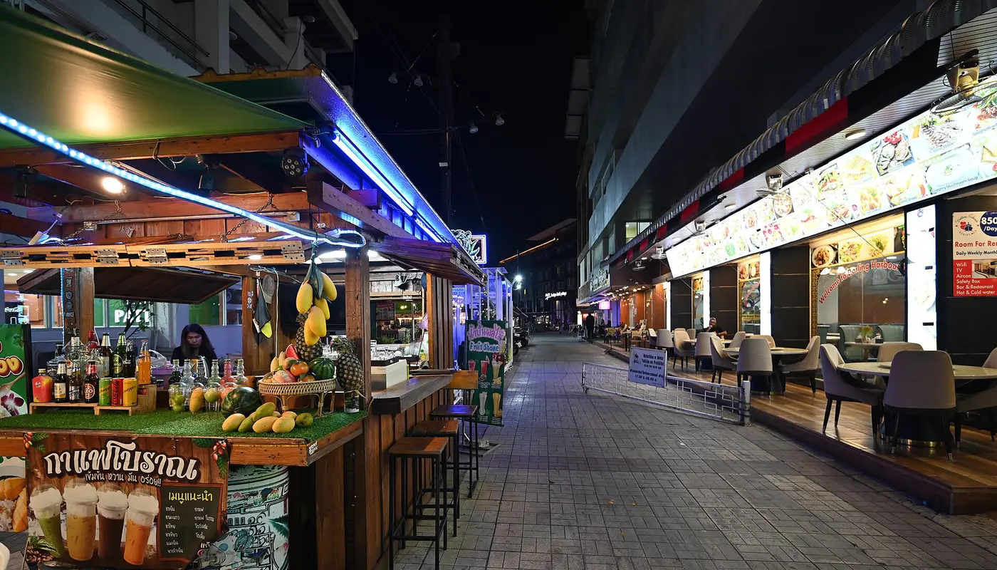 DZ6 1745 Night market alley lined with colorful food stalls and empty sidewalk seating lit by neon signs and warm lanter